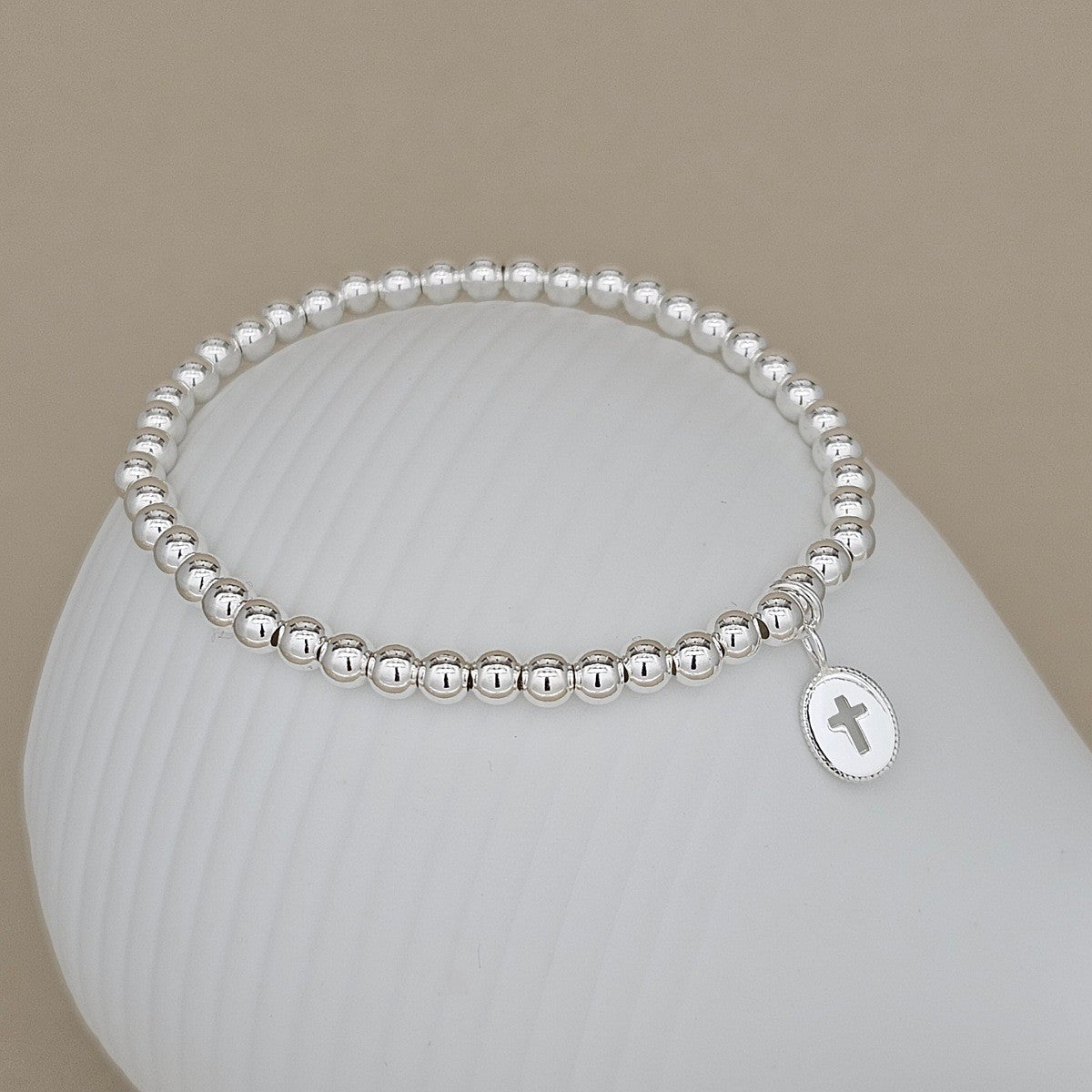 A silver beaded bracelet with a small oval cross pendant, displayed on a white ribbed stand against a beige background.