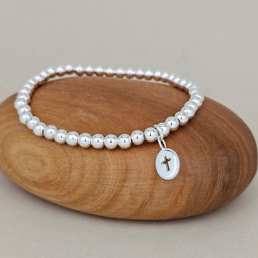 A silver beaded bracelet with a small oval cross pendant, displayed on a smooth brown wooden stone against a neutral beige background.