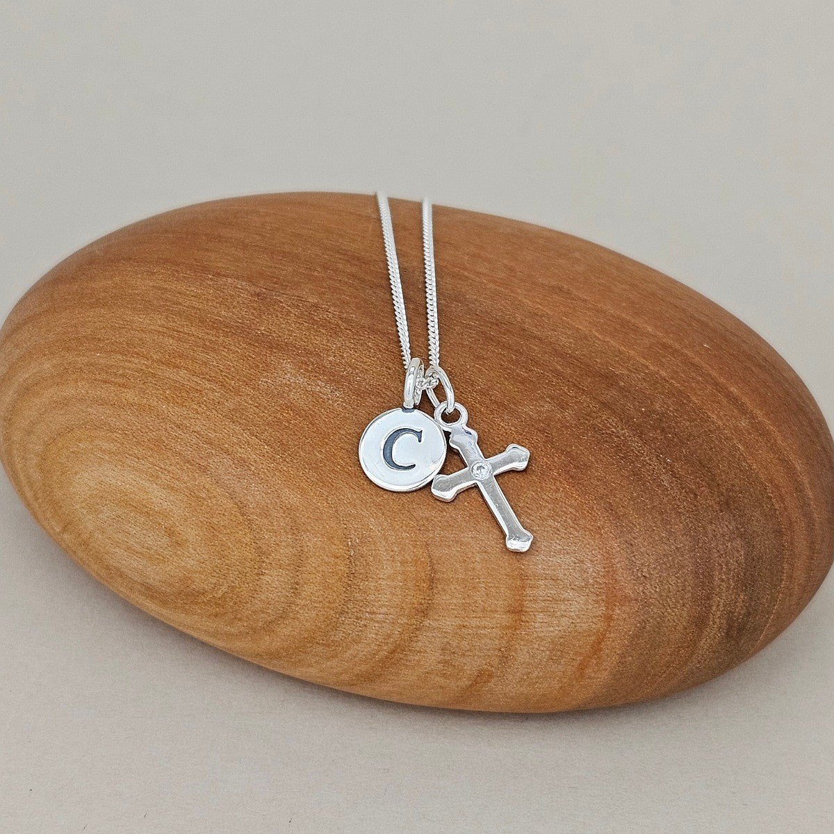 A silver necklace with a round initial 'C' pendant and a small cross pendant, displayed on a smooth, polished wooden stone against a neutral beige background.