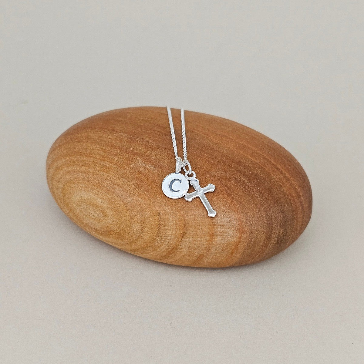 A silver necklace with a small cross and a round initial pendant engraved with the letter 'C', displayed on a smooth, polished wooden stone against a neutral beige background.