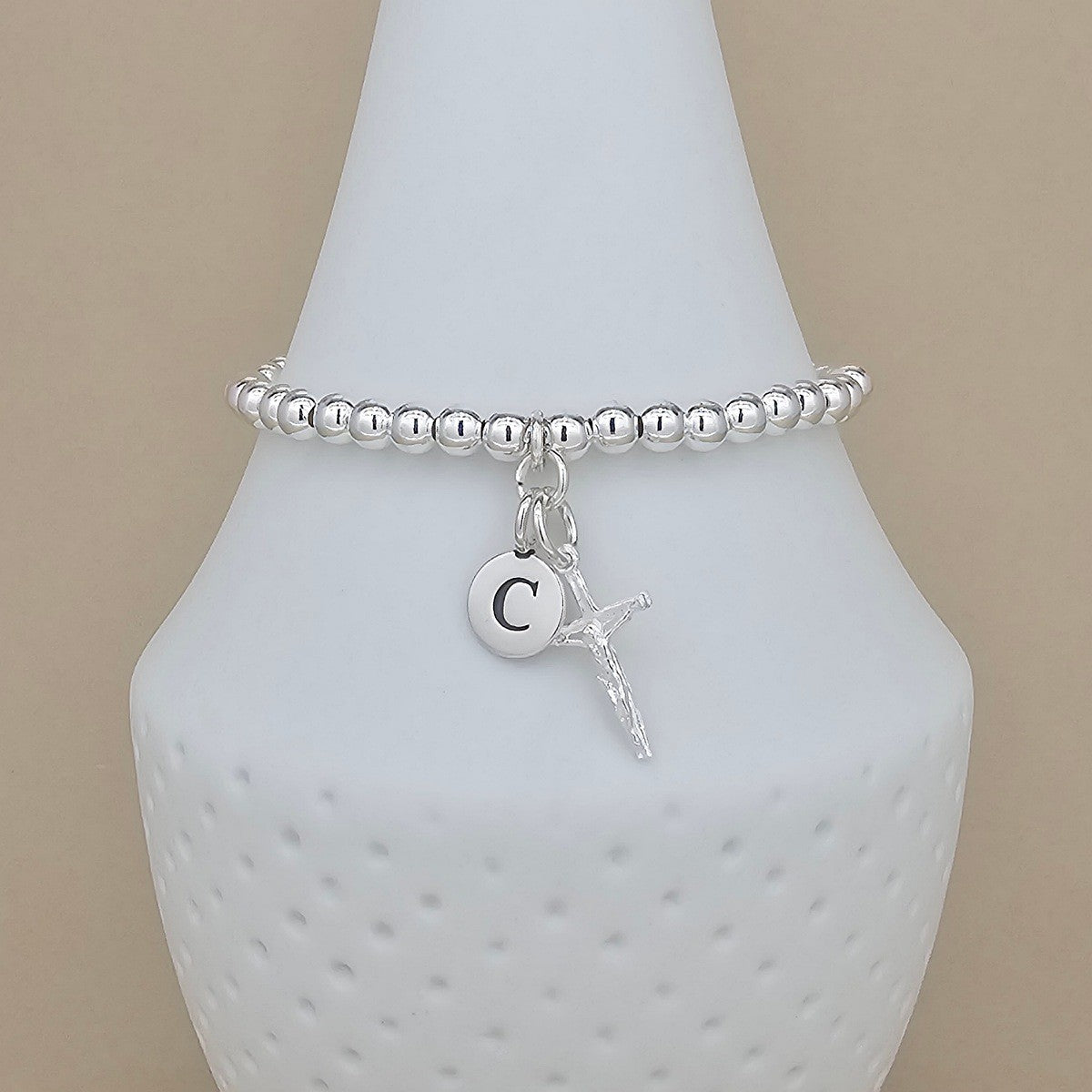 A silver beaded bracelet with a small circular initial 'C' charm and a dangling crucifix, displayed on a white ceramic jewelry bust against a beige background.