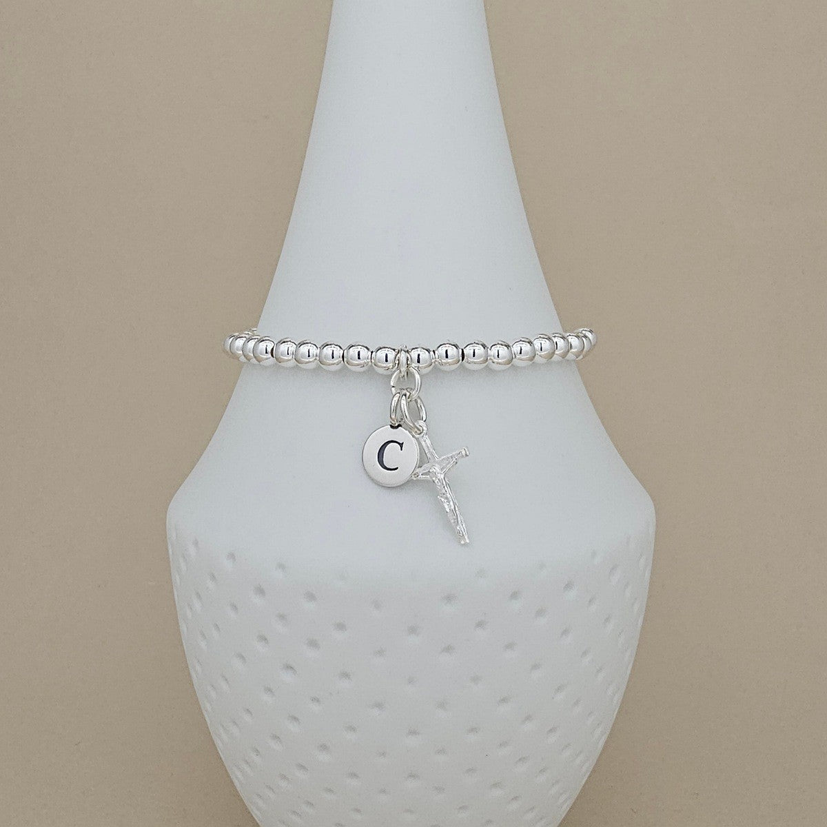 A silver beaded bracelet with a small crucifix charm and a circular initial 'C' pendant, displayed on a white textured cone stand against a beige background.