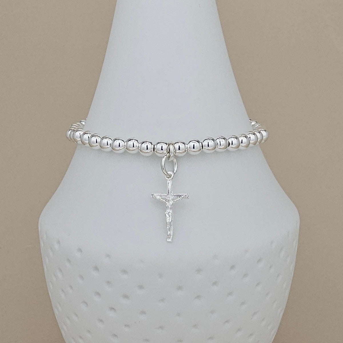 A silver beaded bracelet with a dangling crucifix charm, displayed on a white ceramic jewelry bust against a warm beige background.