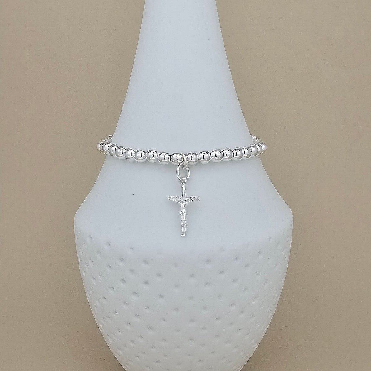 A silver beaded bracelet with a small crucifix charm, displayed on a white textured cone jewelry stand against a warm beige background.