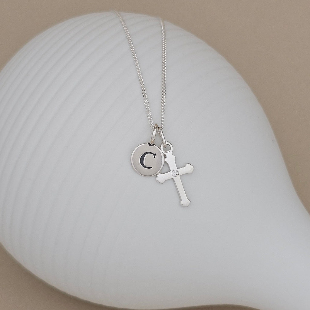 A silver necklace with a small round initial 'C' pendant and a cross charm, displayed on a white ribbed ceramic dish against a beige background.