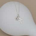 A silver necklace with a small round initial 'C' pendant and a cross charm, displayed on a white ribbed ceramic dish against a beige background.