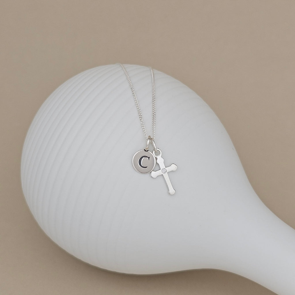 A silver necklace with a small cross pendant and a circular initial 'C' charm, displayed on a white ribbed ceramic stand against a warm beige background.