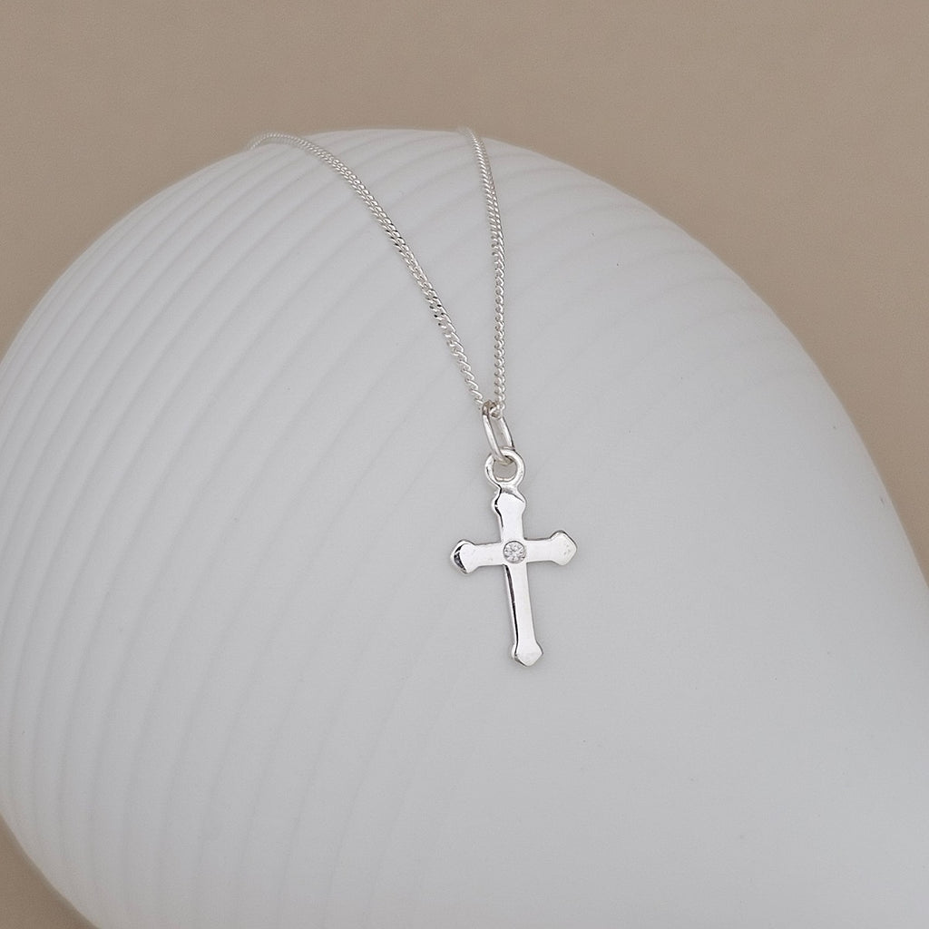 A silver cross pendant necklace with a small clear stone at the center, resting on a white ribbed ceramic surface against a beige background.