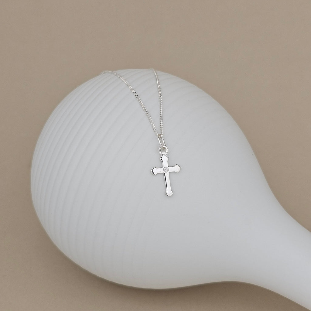 A delicate silver cross pendant necklace on a fine chain, displayed draped over a white ribbed ceramic vase against a solid beige background.