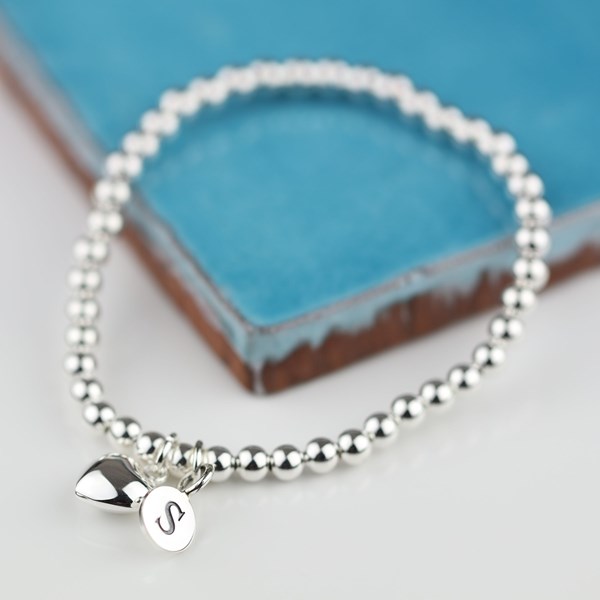 A silver beaded bracelet with a small heart-shaped charm and a round initial 'S' charm, displayed on a light surface with a blue textured book in the background.