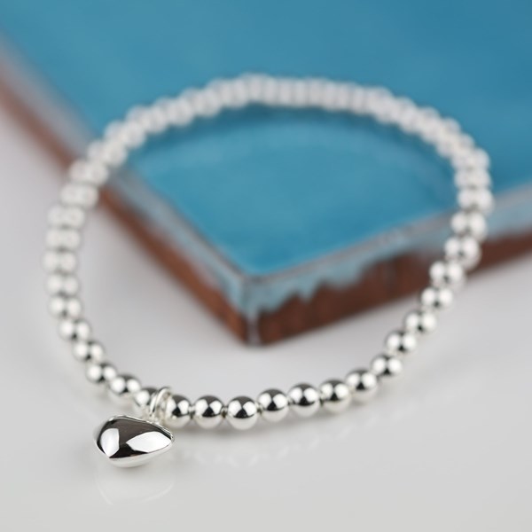 A silver beaded bracelet with a small polished teardrop charm, displayed on a white surface with a blue ceramic tile in the background.