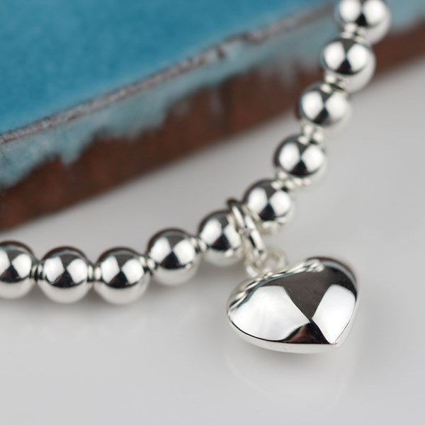 A close-up of a silver beaded bracelet with a polished heart-shaped charm, resting on a white surface with a blue and brown textured background.