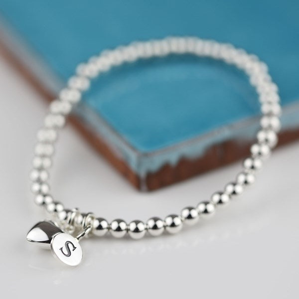 A silver beaded bracelet with a small heart-shaped charm engraved with the letter 'S', displayed on a white surface with a blue ceramic tile in the background.