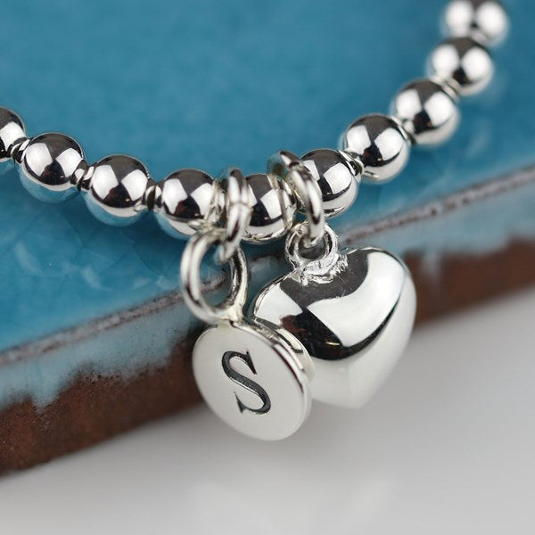 Silver beaded bracelet with heart charm and 'S' initial on a blue and brown background