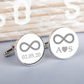 A pair of round silver cufflinks on a light grey surface.  Both cufflinks shows an engraved infinity symbol one with a date below and the other initial letters with an engraved heart symbol in the middle.