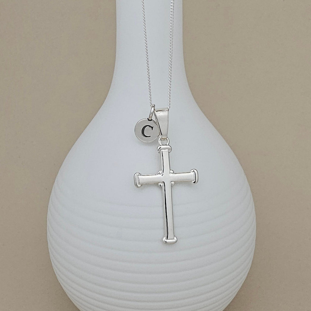 A silver cross pendant necklace with a small circular initial 'C' charm, displayed on a ribbed white vase against a beige background.