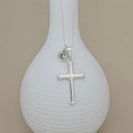 A silver cross pendant necklace with a small circular initial 'C' charm, displayed on a ribbed white vase against a beige background.