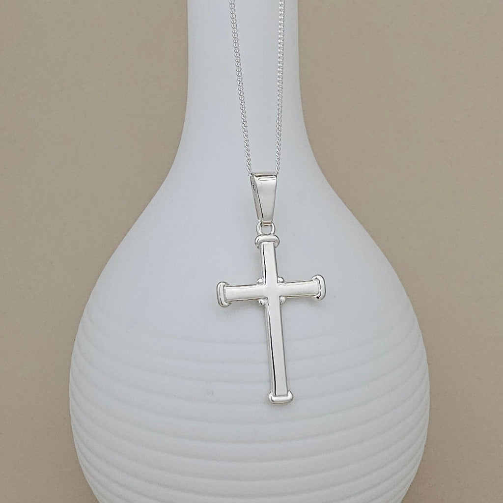 A silver cross pendant necklace displayed on a ribbed white vase against a beige background.