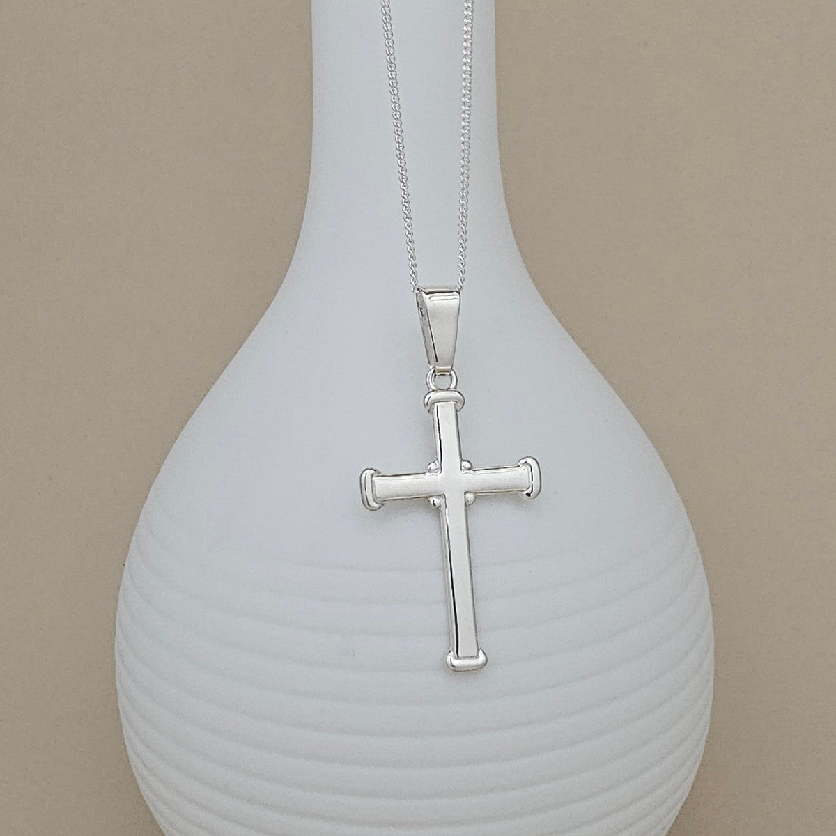 A silver cross pendant necklace displayed on a ribbed white vase against a beige background.