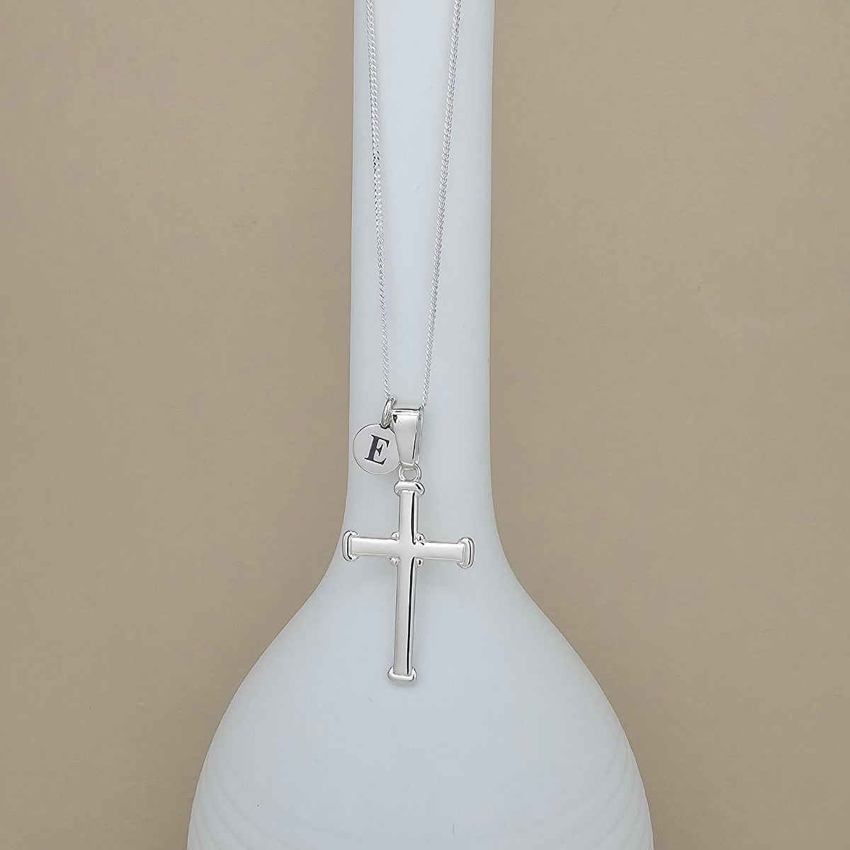 A silver cross pendant necklace with a small initial 'E' charm, displayed on a white ceramic vase against a beige background.