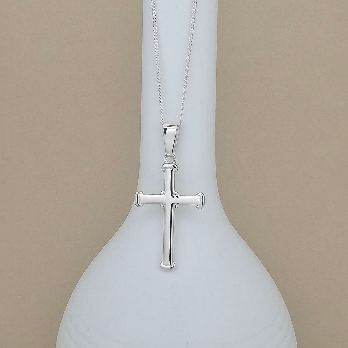 A silver cross pendant necklace hanging from a matching chain, displayed against a white ceramic vase on a warm beige background.