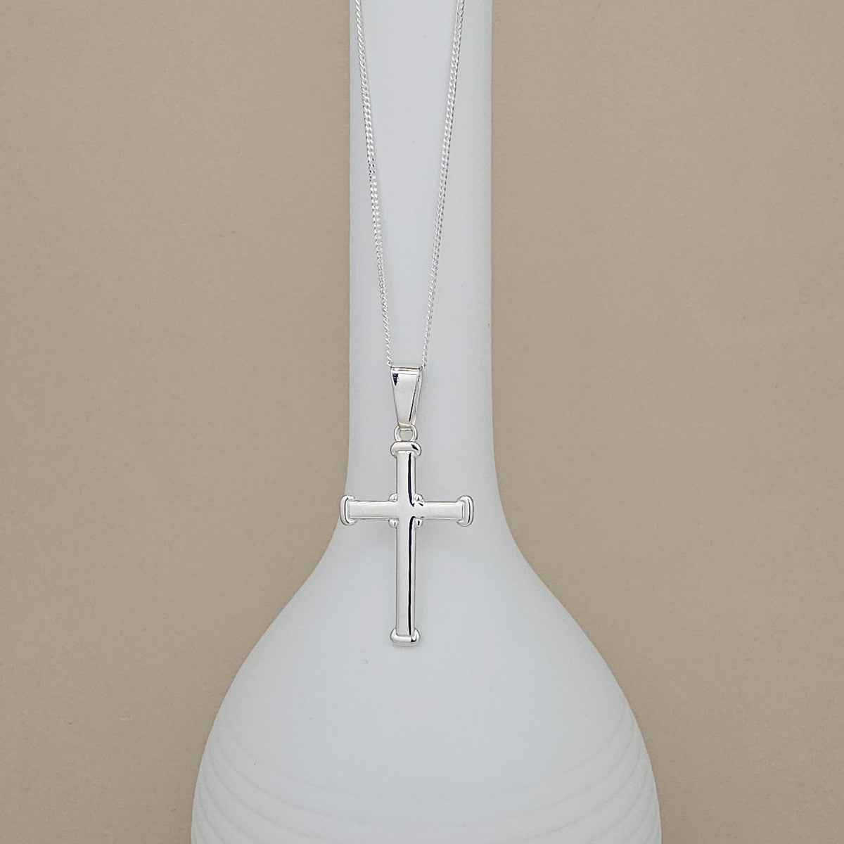 A silver cross pendant necklace hanging from a matching chain, displayed against a white ceramic vase on a warm beige background.