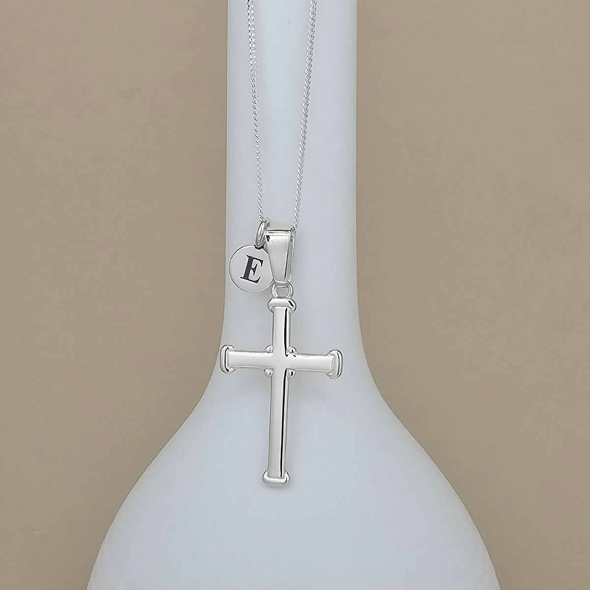 A silver cross pendant necklace with a small round initial 'E' charm, displayed on a white ceramic vase against a beige background.