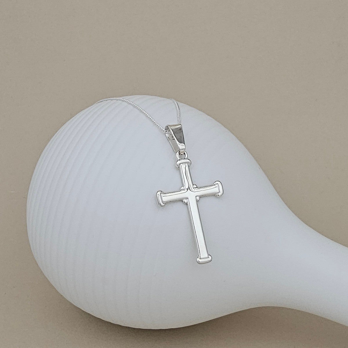 A silver cross pendant necklace on a matching silver chain, displayed on a white ribbed ceramic stand against a solid beige background.