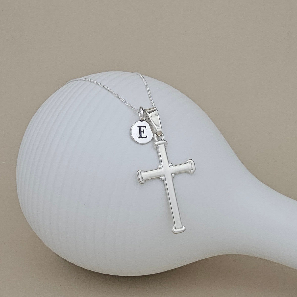 A silver cross pendant necklace with a small round initial 'E' disc charm, displayed on a white ribbed mannequin head against a solid beige background.