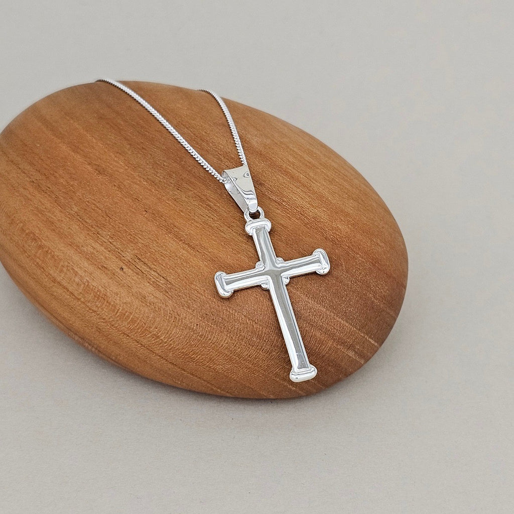 A silver cross pendant necklace on a delicate silver chain, displayed on a smooth, polished wooden stone against a neutral gray background.