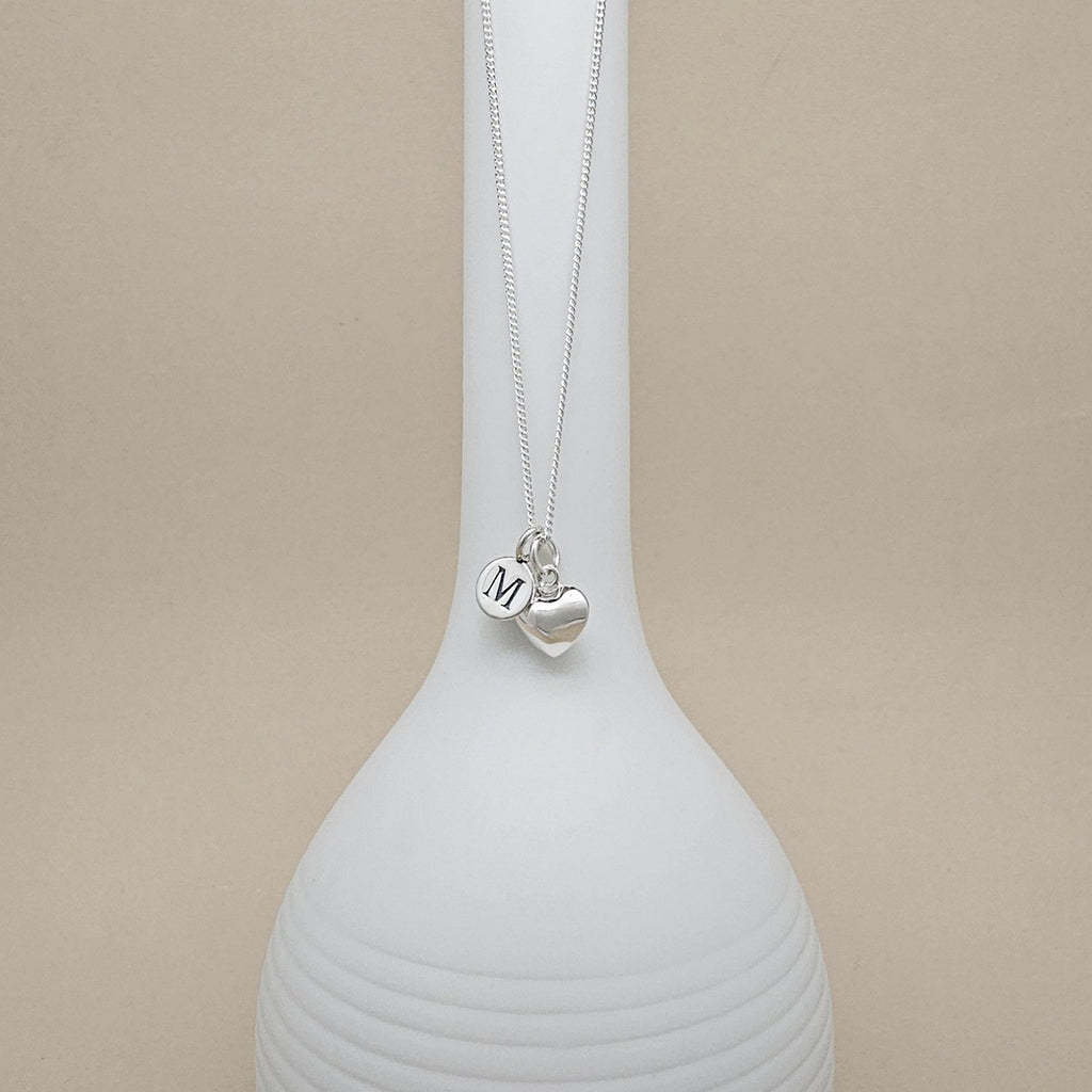 A silver necklace with heart-shaped charm and engraved charm with the letter 'M', displayed on a white ceramic vase against a beige background.