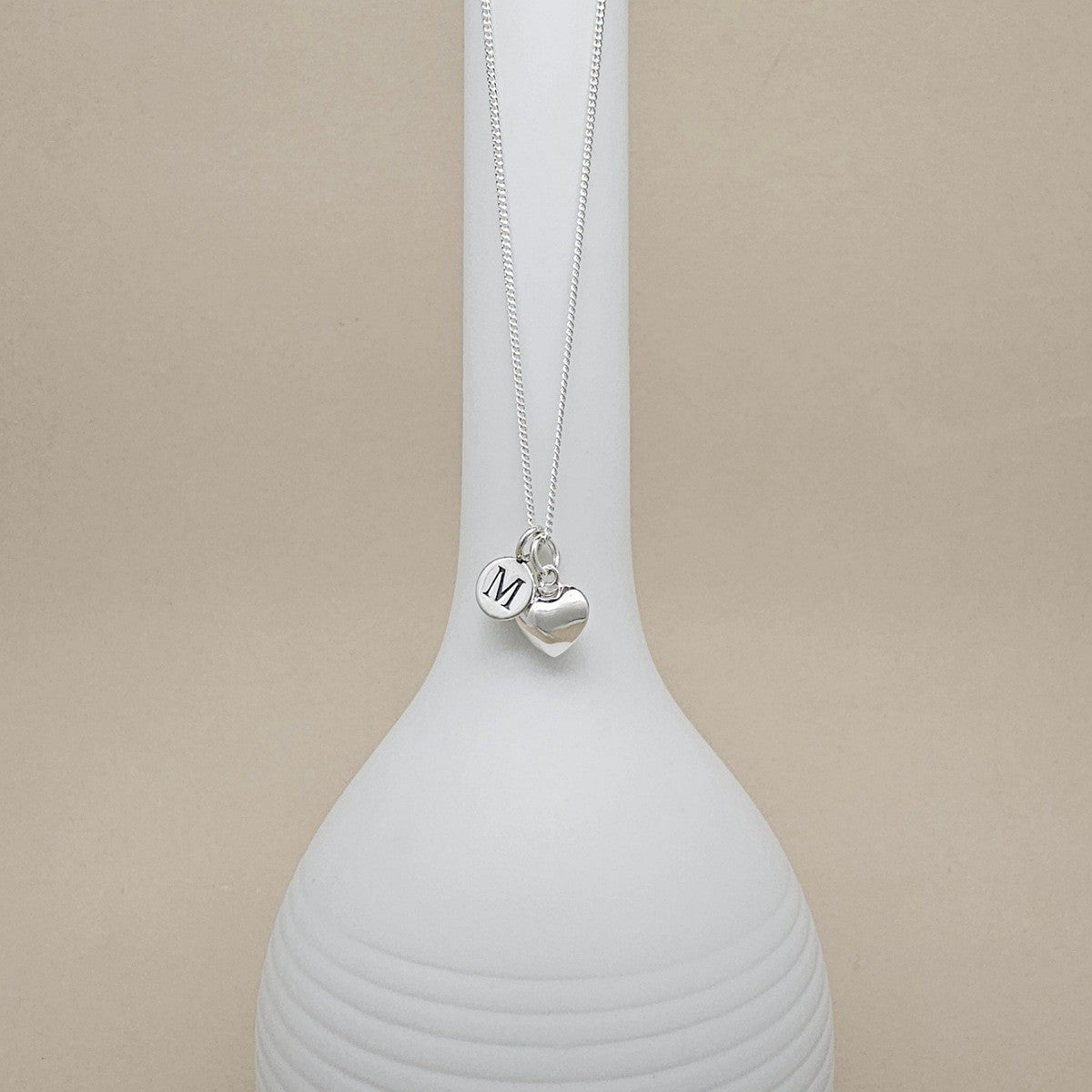 A silver necklace with heart-shaped charm and engraved charm with the letter 'M', displayed on a white ceramic vase against a beige background.