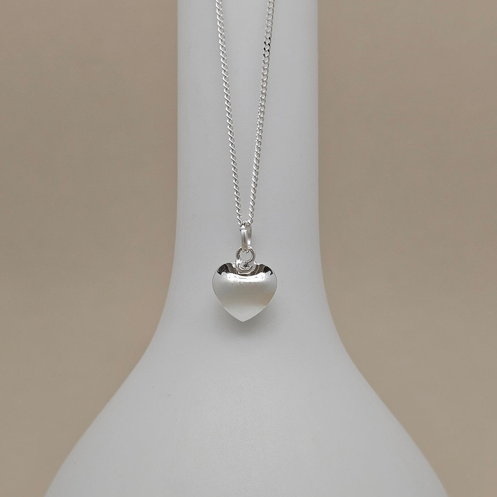 A silver heart pendant necklace displayed on a white neck mannequin against a warm beige background.