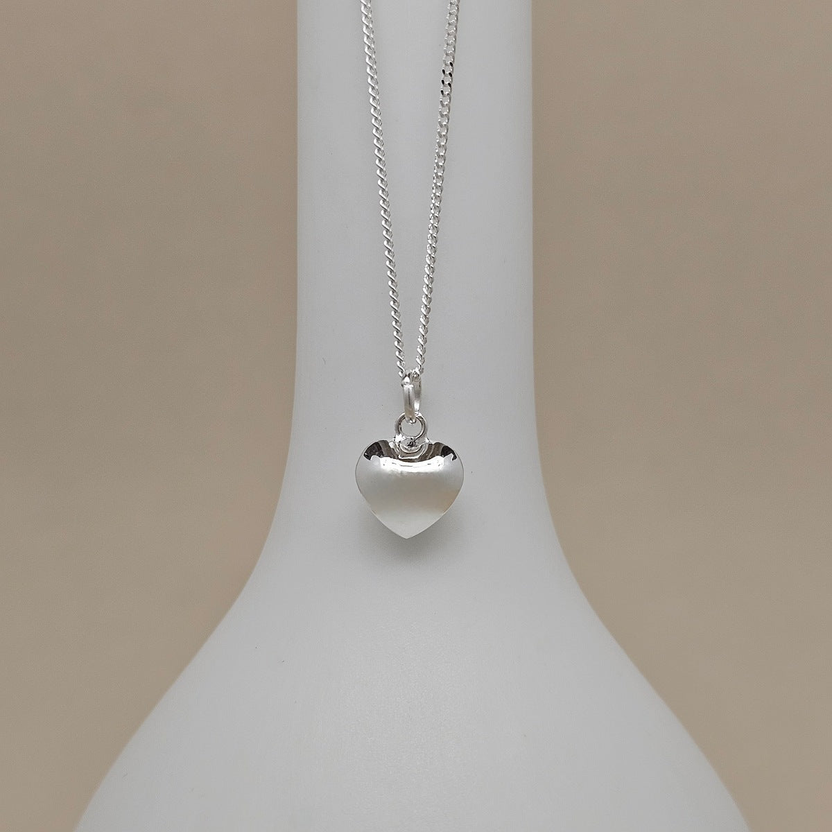 A silver heart pendant necklace displayed on a white neck mannequin against a warm beige background.