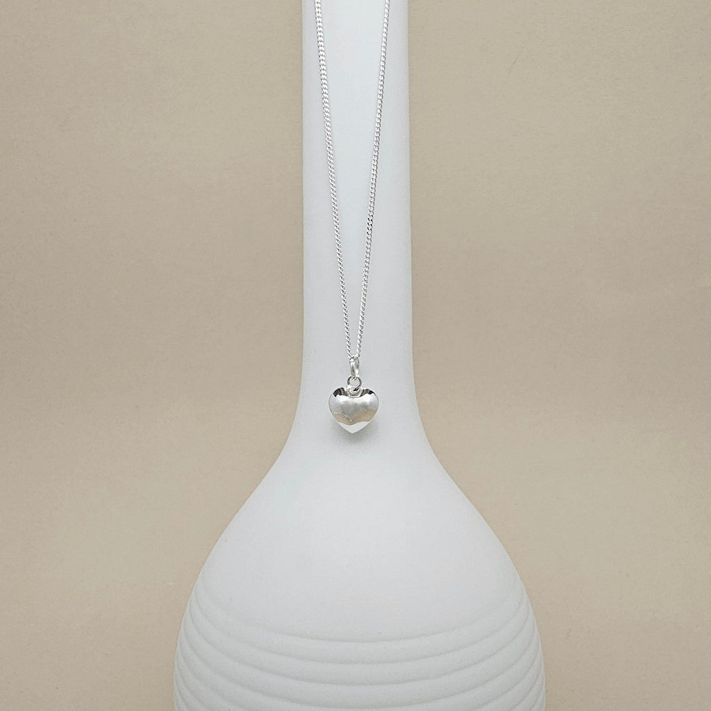 A silver heart pendant necklace on a delicate chain, displayed on a white ceramic vase against a warm beige background.