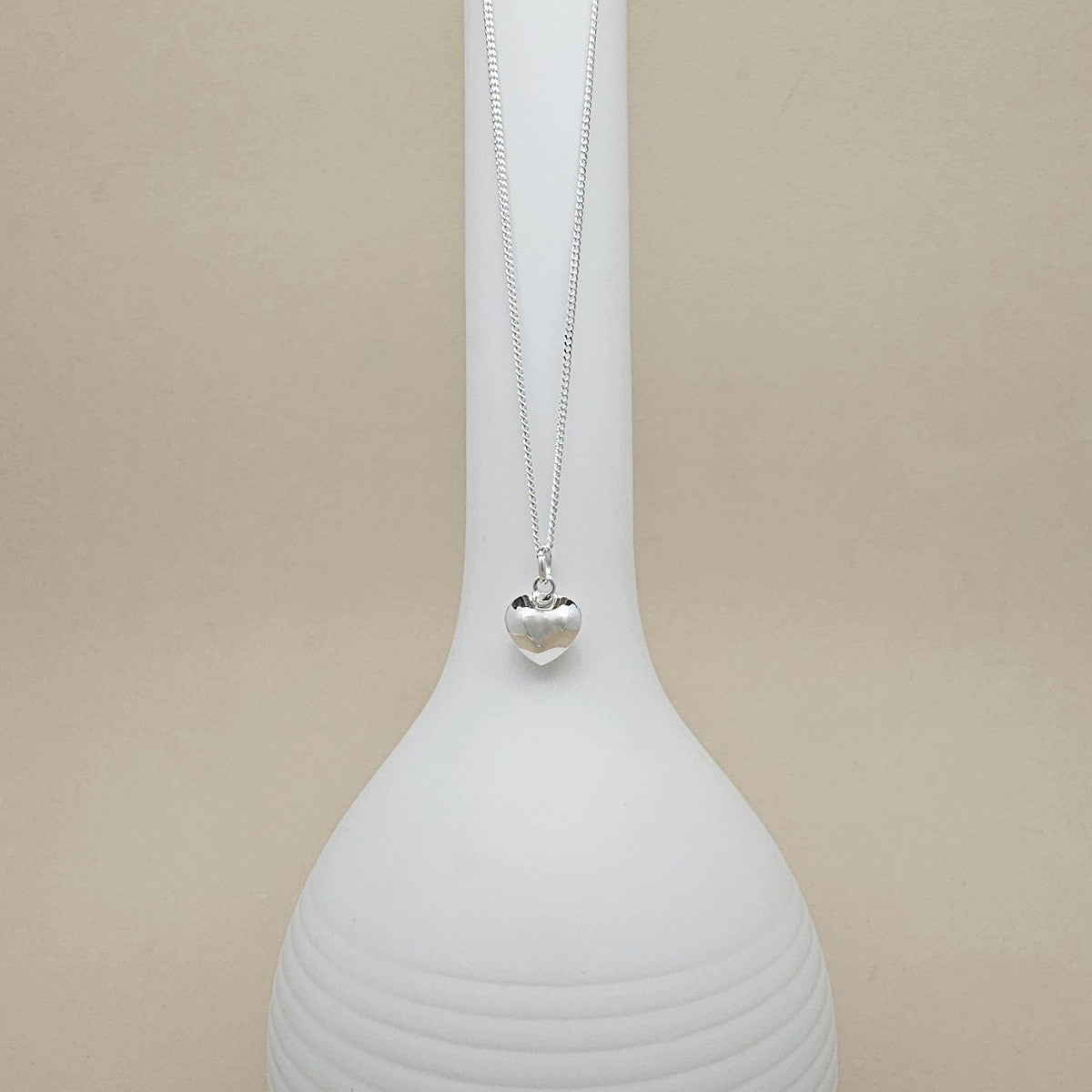 A silver heart pendant necklace on a delicate chain, displayed on a white ceramic vase against a warm beige background.