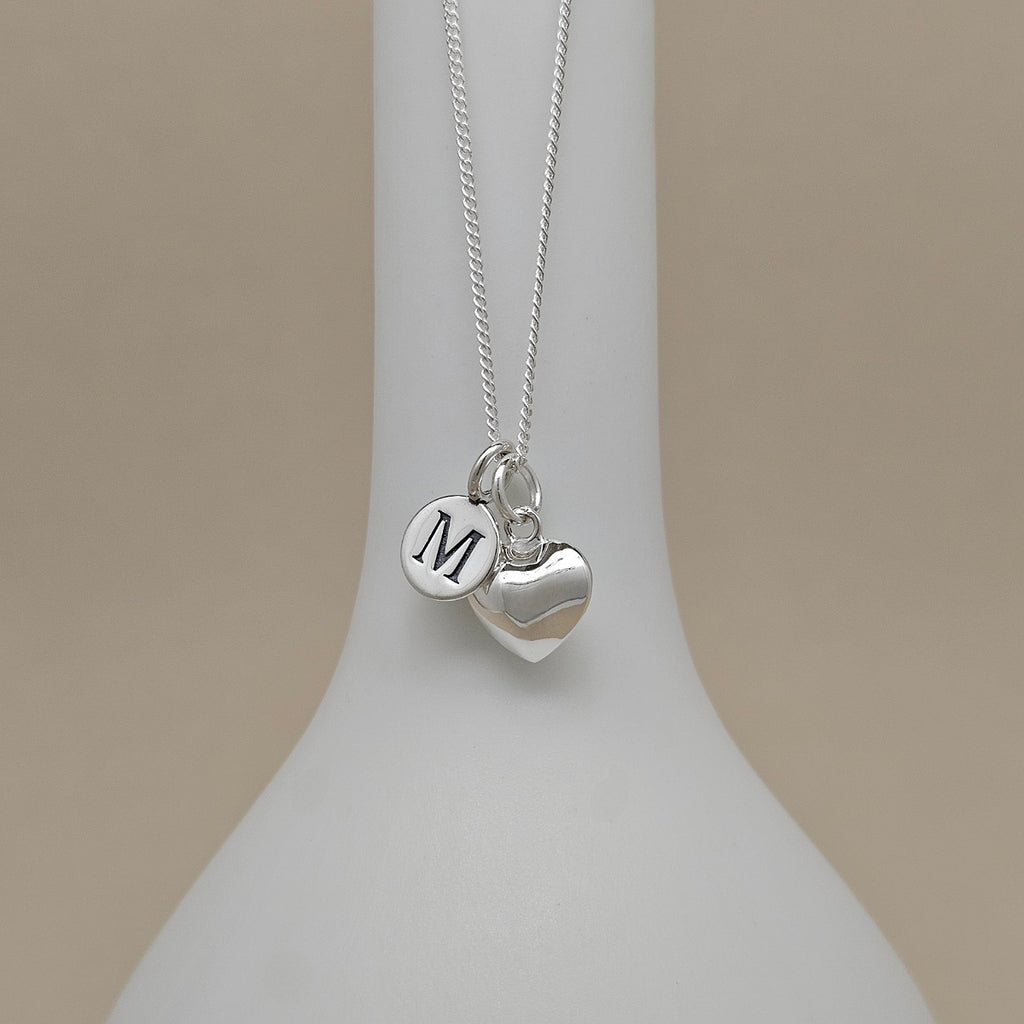 A silver necklace with a heart-shaped charm, with an engraved charm with the letter 'M', displayed on a white jewelry bust against a warm beige background.