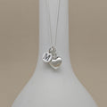 A silver necklace with a heart-shaped charm, with an engraved charm with the letter 'M', displayed on a white jewelry bust against a warm beige background.