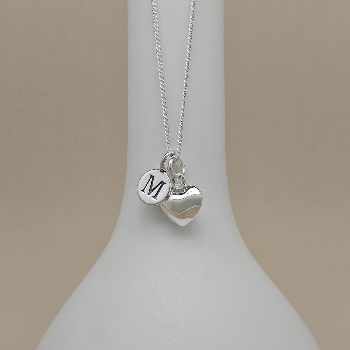 A silver necklace with a heart-shaped charm, with an engraved charm with the letter 'M', displayed on a white jewelry bust against a warm beige background.