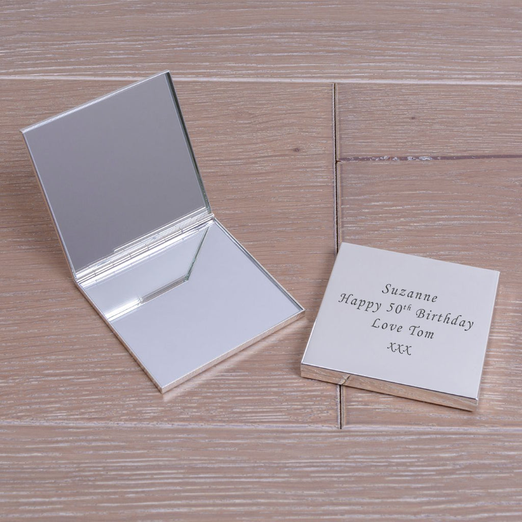 Two silver square compact mirrors one displayed open with two internal mirrors,  the other closed showing the engraved wording 'Suzanne Happy 50th Birthday Love Tom xxx' displayed on a wooden surface.