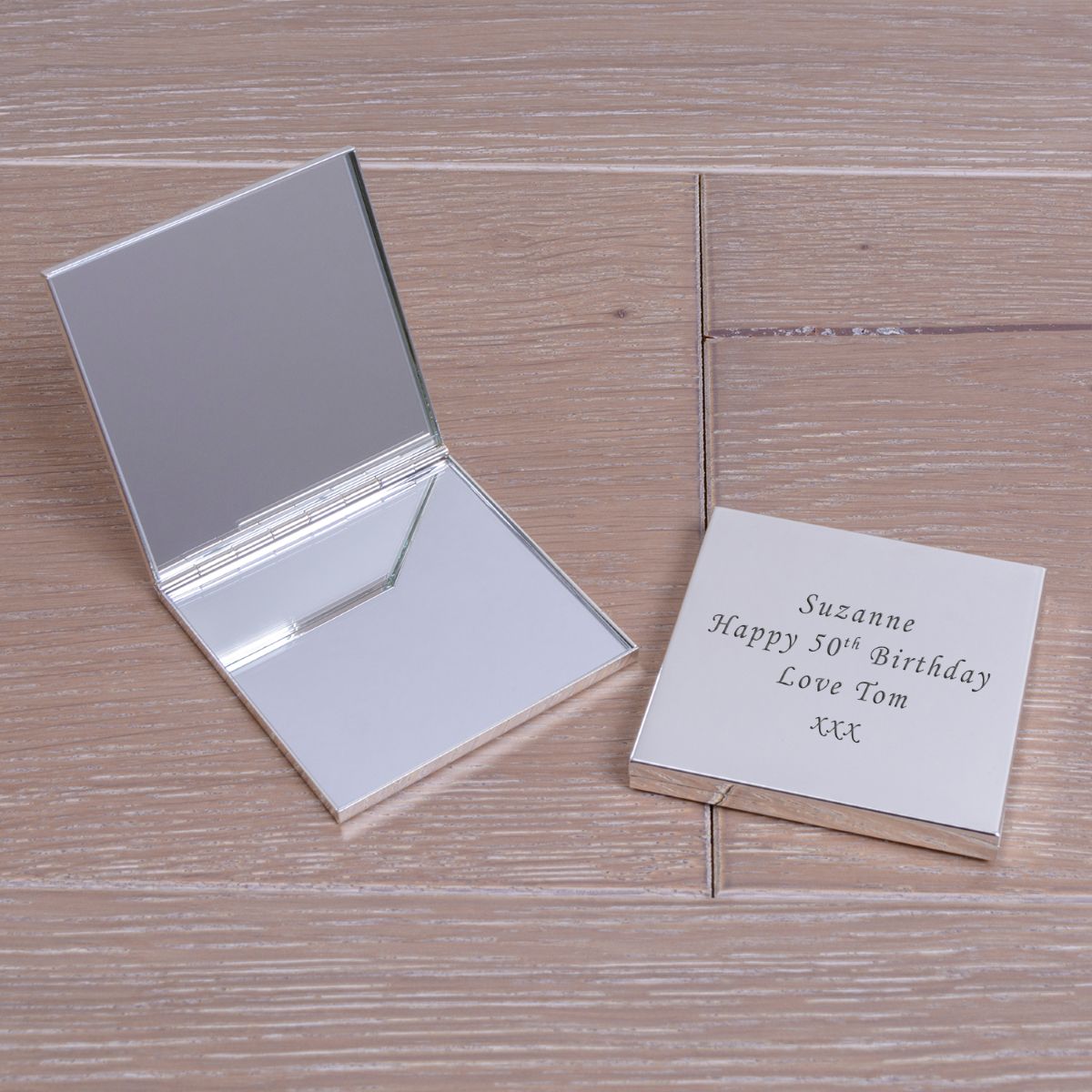 Two silver square compact mirrors one displayed open with two internal mirrors,  the other closed showing the engraved wording 'Suzanne Happy 50th Birthday Love Tom xxx' displayed on a wooden surface.