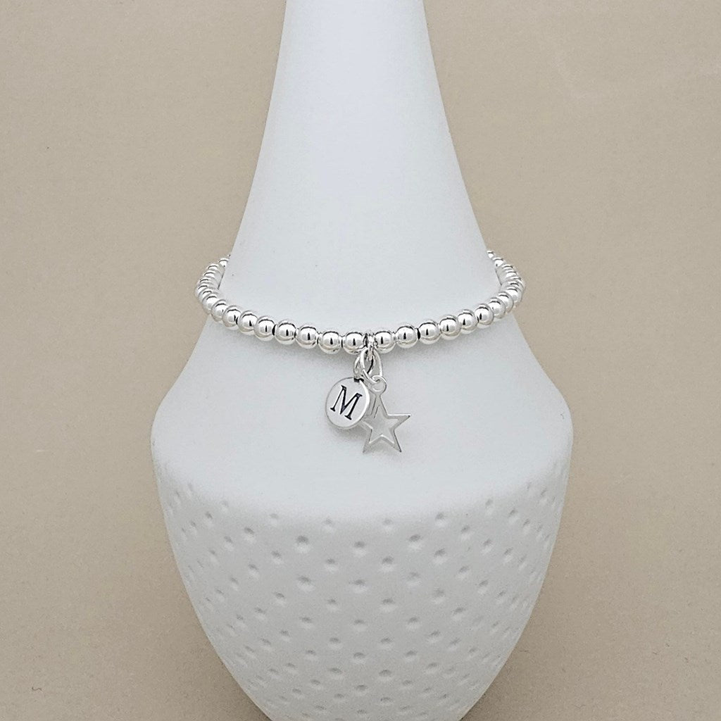 A silver beaded bracelet with a round initial 'M' charm and a star charm, displayed on a white textured cone stand against a beige background.