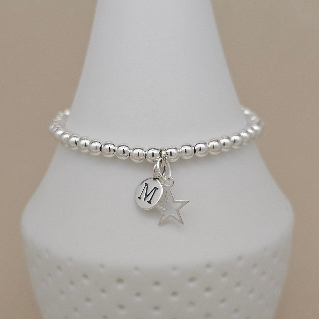 A silver beaded bracelet with a circular initial 'M' charm and a star charm, displayed on a white cone stand against a beige background.