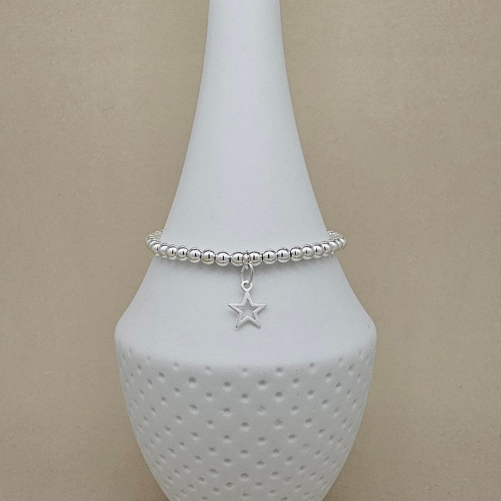 A silver beaded bracelet with a dangling star charm, displayed on a white ceramic jewelry stand against a beige background.