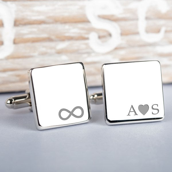 A pair of square silver cufflinks on a light grey surface. one cufflink shows an engraved infinity symbol and the other initial letters with an engraved heart symbol in the middle.