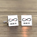 A pair of square silver-toned cufflinks on a light wood surface. Both cufflink faces are white with a black infinity symbol with small wording 'You Me' in red within the symbol and a printed date underneath.