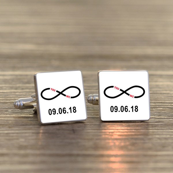 A pair of square silver-toned cufflinks on a light wood surface. Both cufflink faces are white with a black infinity symbol with small wording 'You Me' in red within the symbol and a printed date underneath.