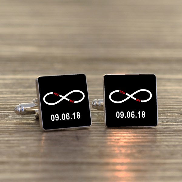 A pair of square silver-toned cufflinks on a light wood surface. Both cufflink faces are black with a white infinity symbol with small wording 'You Me' in red within the symbol and a printed date underneath.