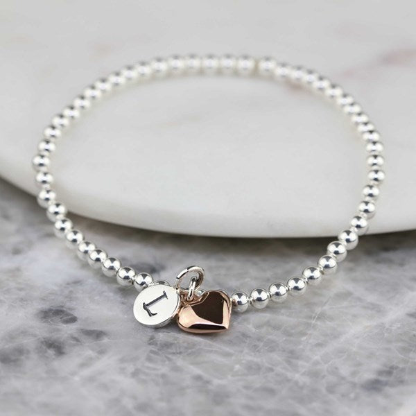 A silver beaded bracelet with two heart charms, one polished silver and one rose gold, displayed on a white stone slab over a gray marble surface.