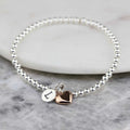 A silver beaded bracelet with two heart charms, one polished silver and one rose gold, displayed on a white stone slab over a gray marble surface.
